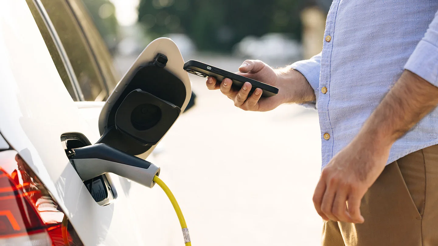Ein Mann schließt ein Ladekabel an ein Elektroauto an und hat ein Smartphone in der Hand