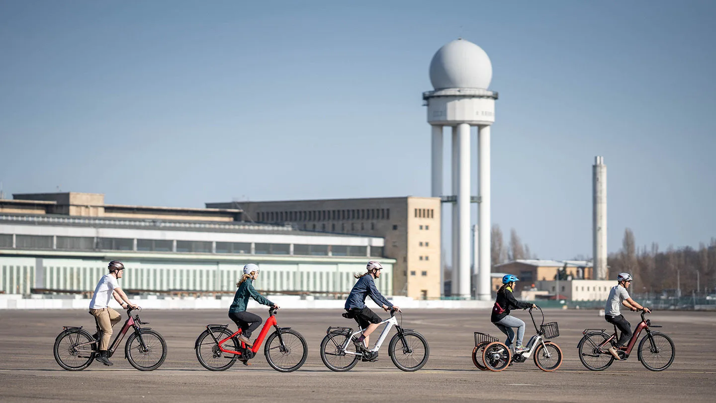 Fünf Personen fahren auf unterschiedlichen E-Bikes und einem E-Dreirad über einen weitläufigen Platz vor dem ehemaligen Flughafen Berlin-Tempelhof