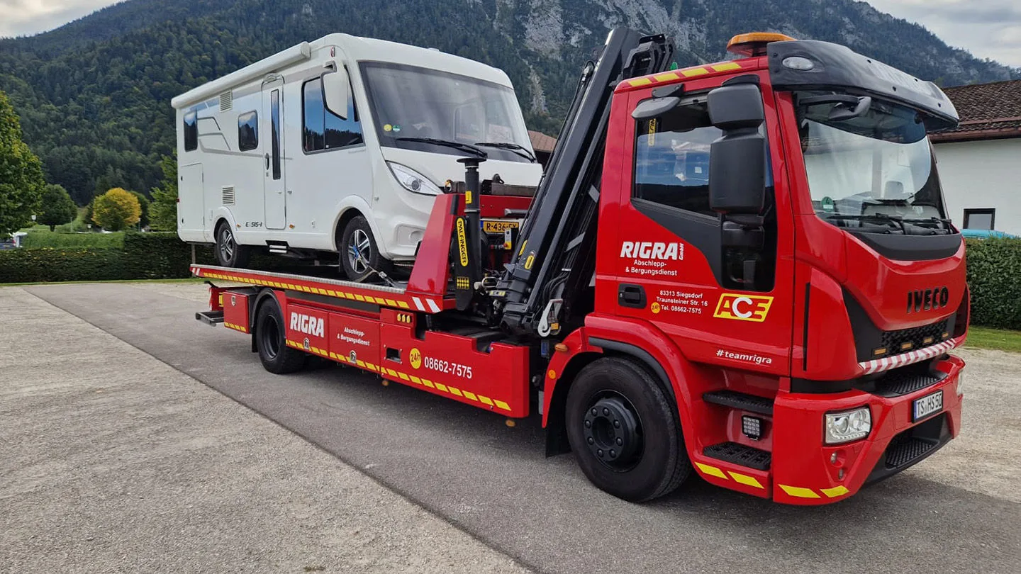 Das Bild zeigt einen roten ACE-Abschleppwagen, der ein weißes Wohnmobil auf seiner Ladefläche transportiert.