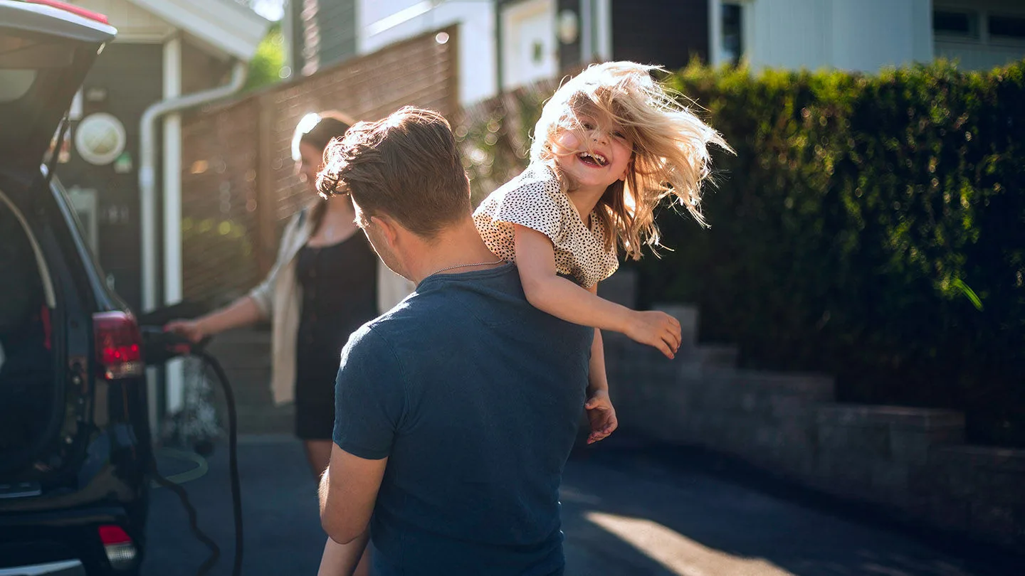 Ein Vater spielt mit seiner Tochter während die Mutter das E-Auto der Familie lädt.