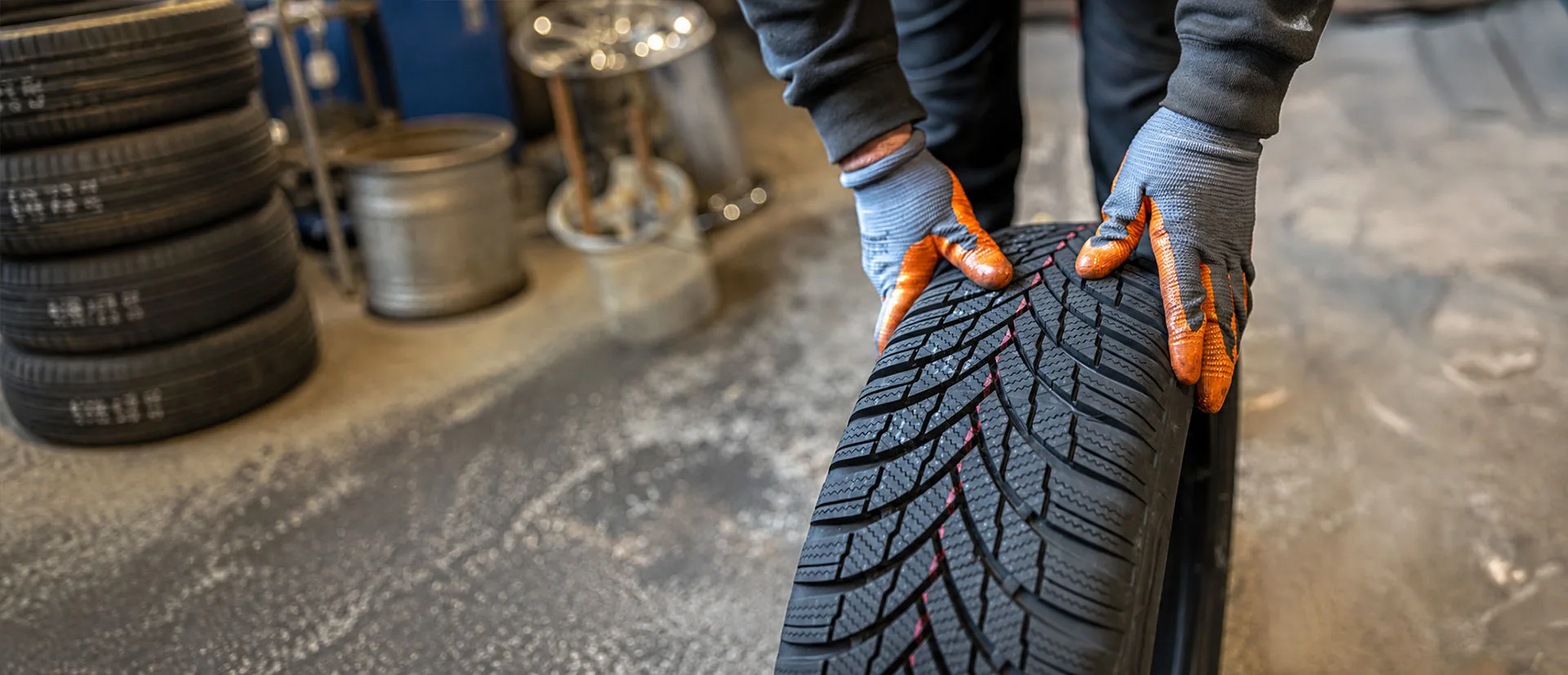 Mechaniker mit Arbeitshandschuhen hält einen neuen Autoreifen in einer Werkstatt; im Hintergrund sind weitere Reifen und Felgen zu sehen.