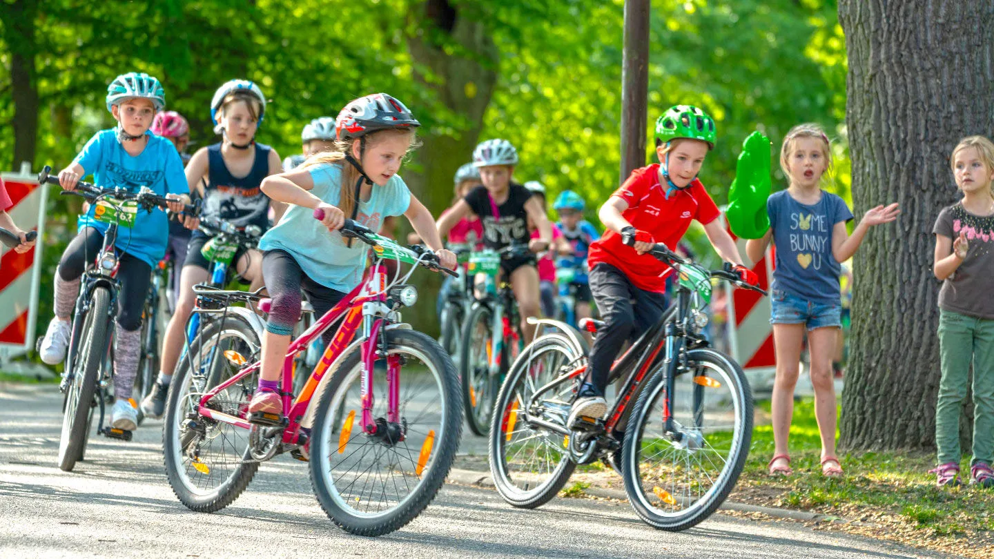 Mehrere Kinder fahren mit Helmen auf Fahrrädern bei einer Veranstaltung oder einem Fahrradrennen auf einem abgesperrten Weg. Am Rand stehen weitere Kinder und schauen zu.