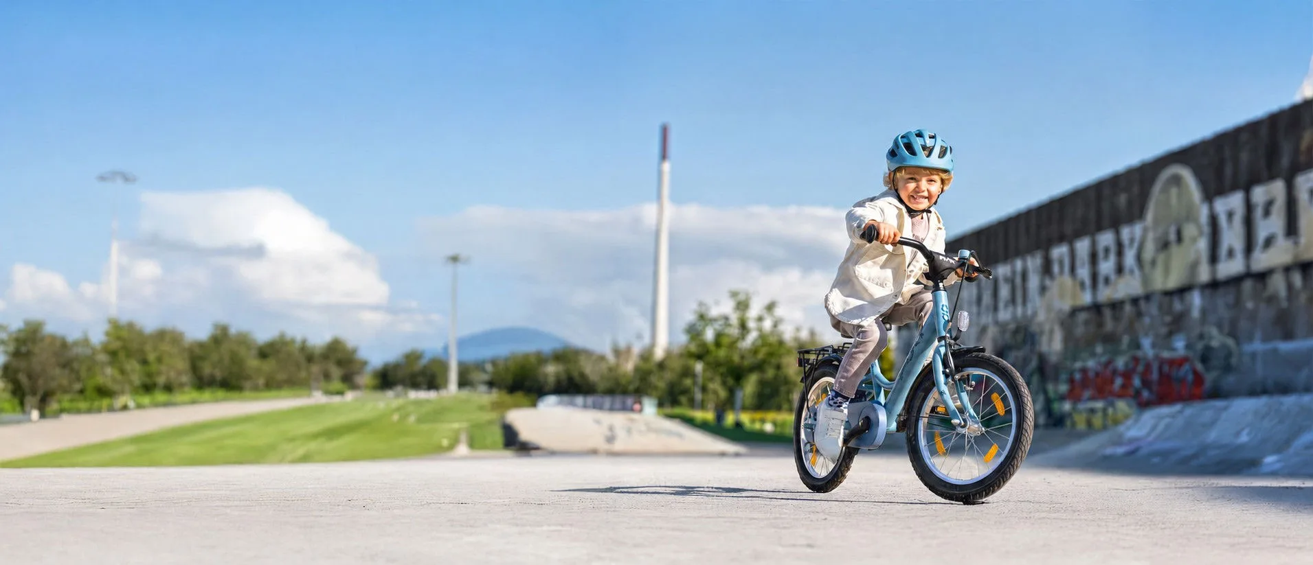 Ein Kind fährt mit Helm auf einem Kinderfahrrad durch einen Skatepark. Im Hintergrund sind eine Graffitiwand, Grünflächen und ein sonniger Himmel zu sehen.