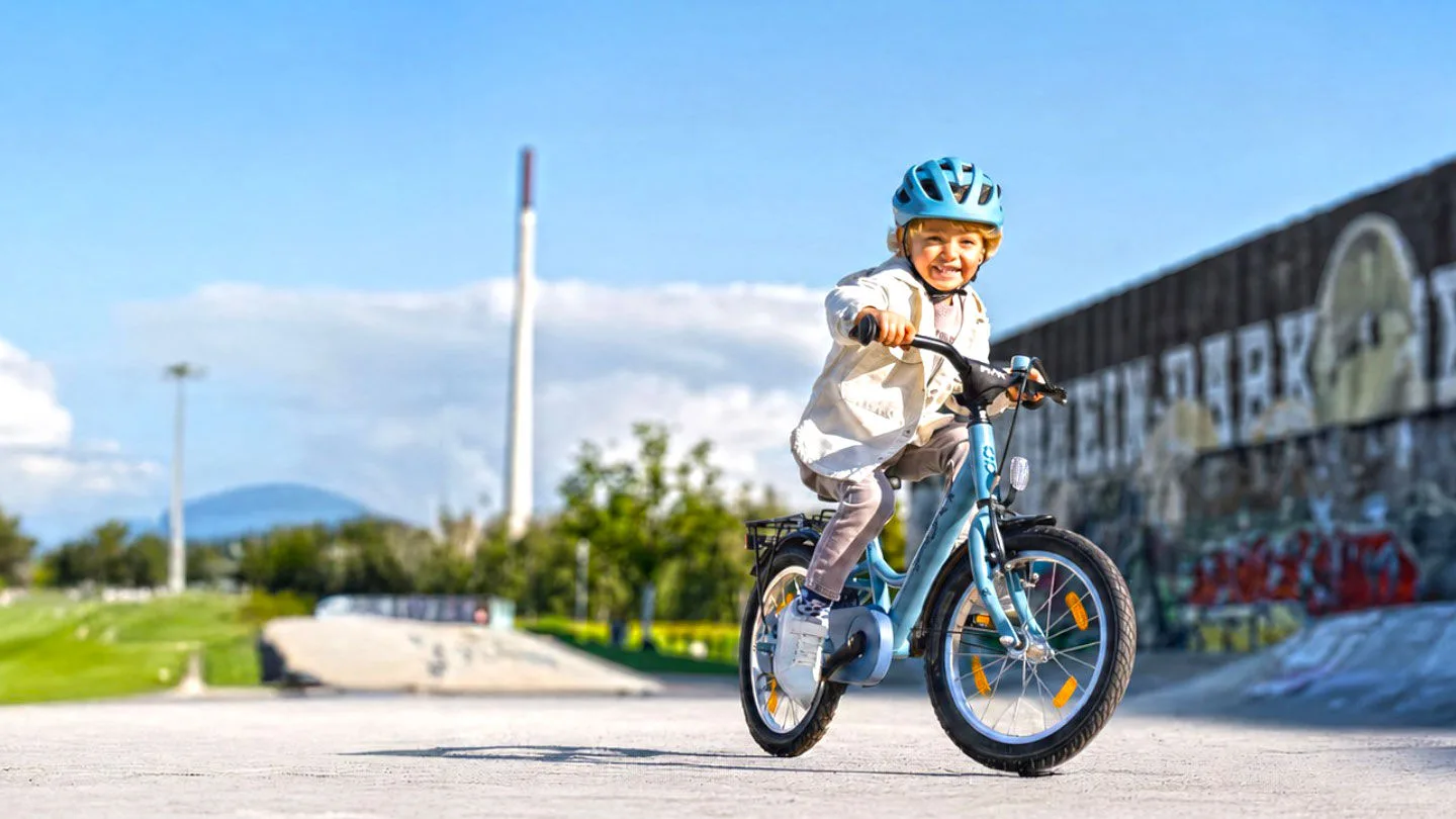Ein Kind fährt mit Helm auf einem Kinderfahrrad durch einen Skatepark. Im Hintergrund sind eine Graffitiwand, Grünflächen und ein sonniger Himmel zu sehen.