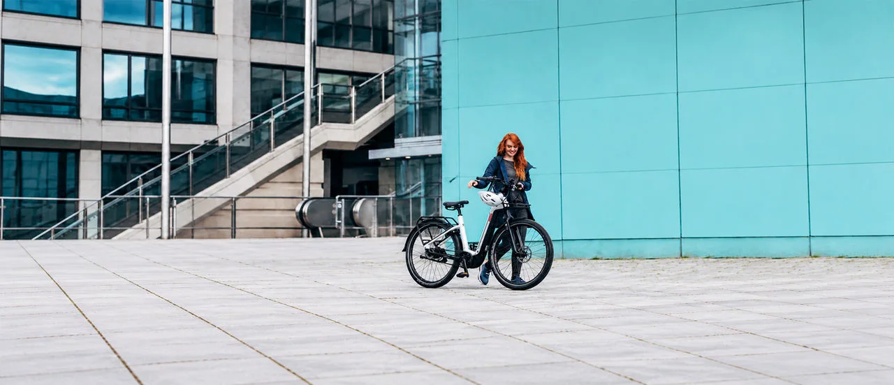 Frau mit roten Haaren steht mit einem schwarzen E-Bike auf einem weitläufigen Platz vor einem modernen Gebäude mit türkisfarbener Fassade