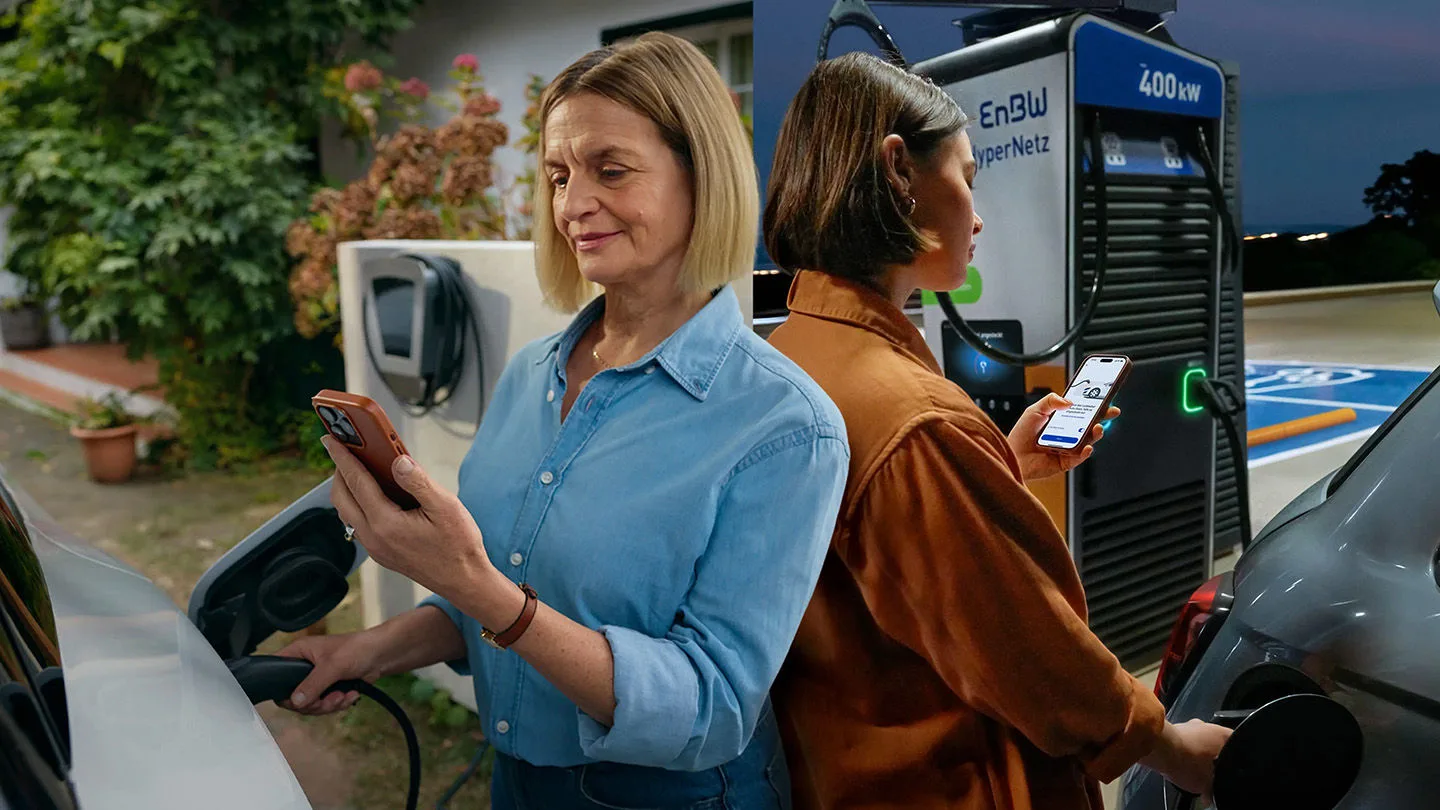 Zwei Frauen stehen Rücken an Rücken und laden ihre Elektroautos an einer EnBW Ladesäule. Dabei schauen sie auf ihren Smartphones auf die EnBW App.