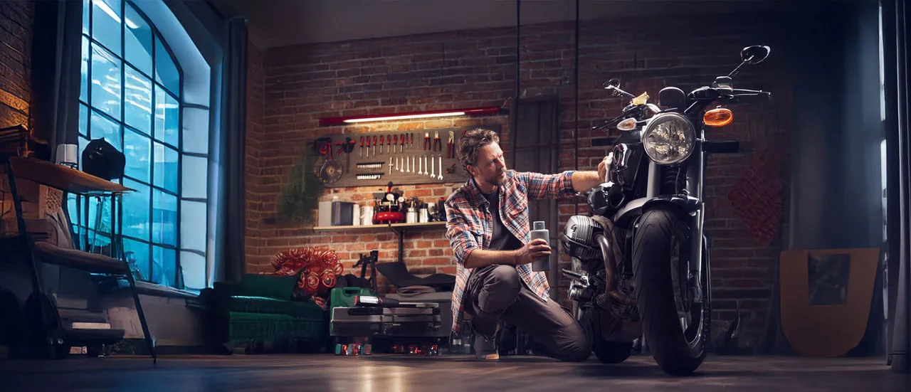 Mann poliert sein vintage Motorrad in seiner Garage, er hält eine Flasche Wachs in der einen Hand und bringt es mit der anderen Hand mit einem Tuch auf.