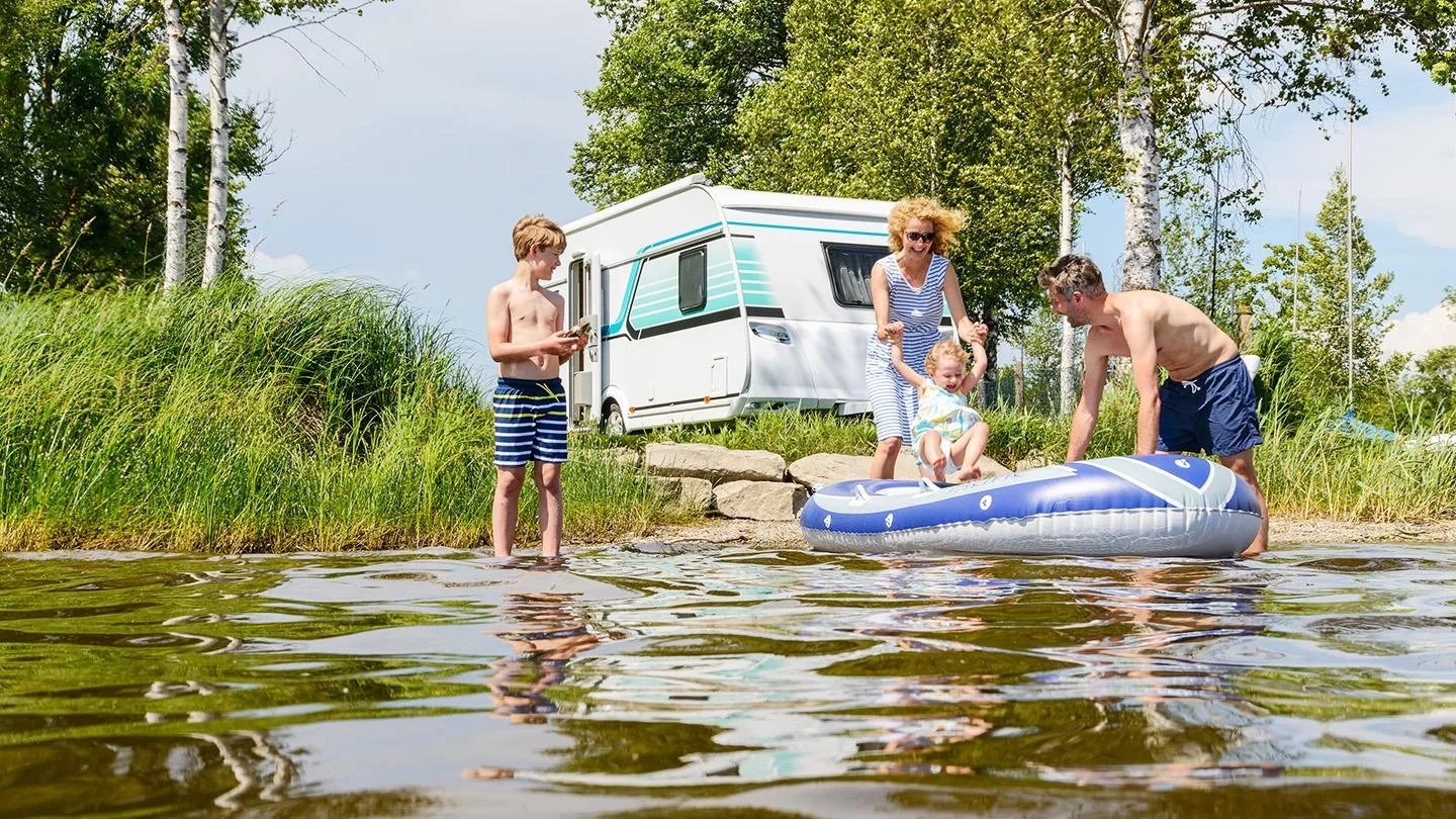 Familie schwimmt in einem See im Hintergrund steht ein Wohnmobil 