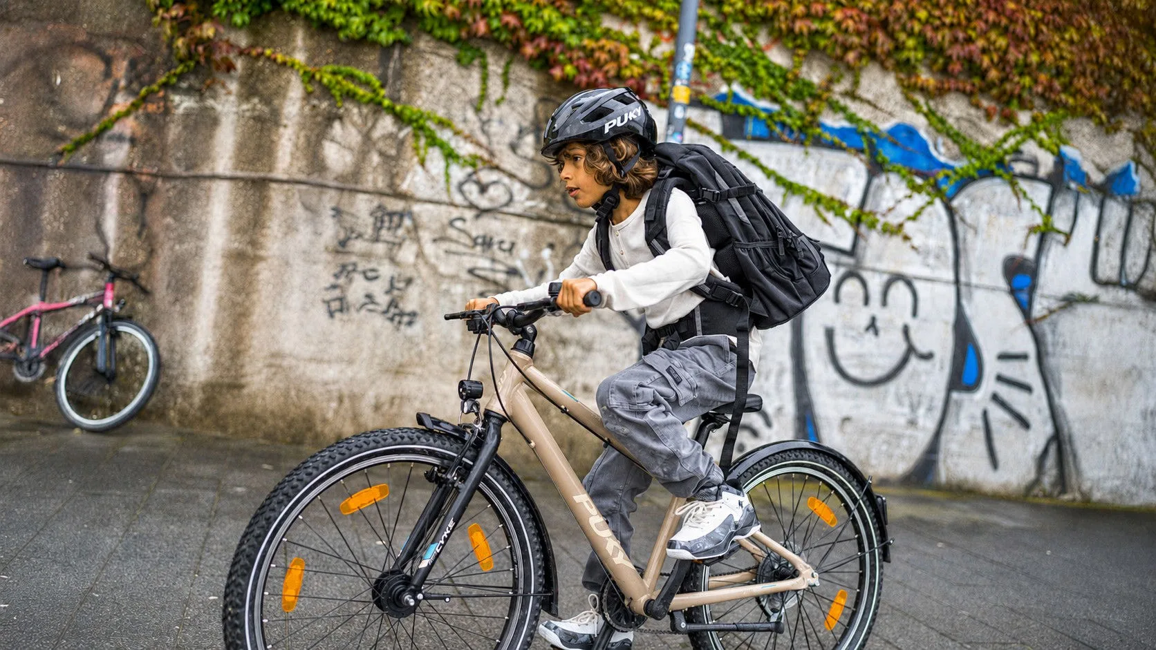 Ein Kind fährt mit Helm und Rucksack auf einem Kinderfahrrad an einer Graffitiwand entlang. Das Fahrrad ist mit Reflektoren, Beleuchtung und Schutzblechen ausgestattet.